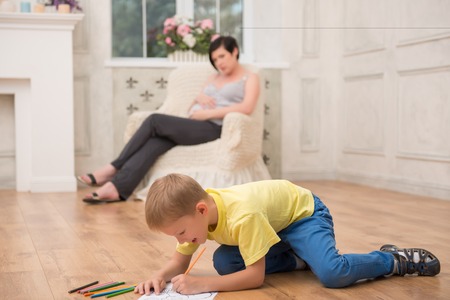 Selected focus on little fair-haired smiling boy wearing yellow T-shirt and jeans drawing nice picture for his pregnant mom sitting in the comfortable chair on backgroundの写真素材