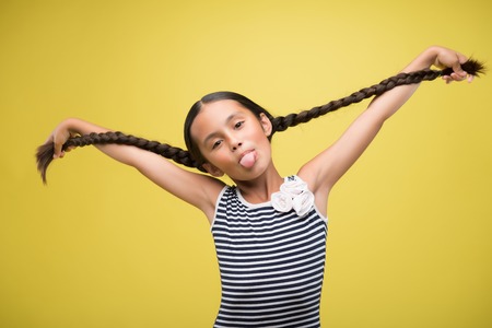Half-length portrait of pretty dark-haired funny little girl wearing nice striped dress with white rose on it showing us her tongue and keeping at her plaits. Isolated on yellow background の写真素材