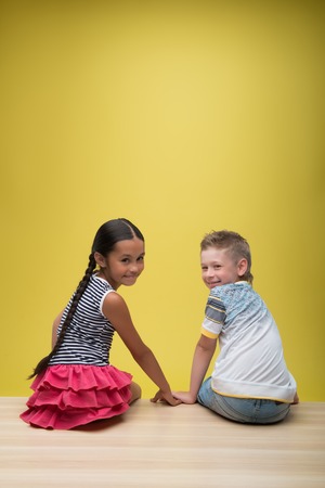 Half-length portrait of pretty dark-haired smiling little girl wearing nice striped dress sitting back with little fair-haired smiling boy both looking at us. Isolated on yellow background の写真素材