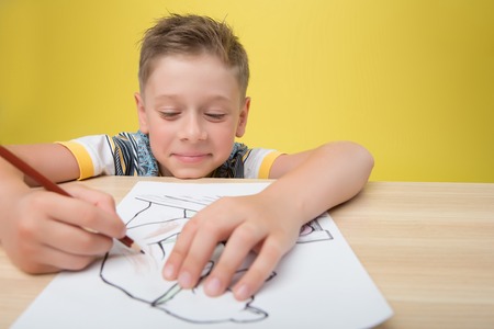 Little smiling fair-haired funny boy wearing nice colored T-shirt sitting at the table drawing a picture. Isolated on yellow backgroundの写真素材