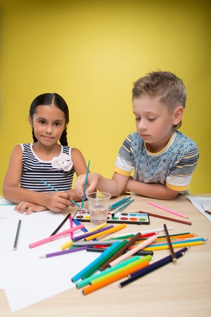 Half-length portrait of pretty dark-haired little girl wearing nice striped dress sitting at the table with many watercolors on it painting with little fair-haired boy. Isolated on yellow background の写真素材