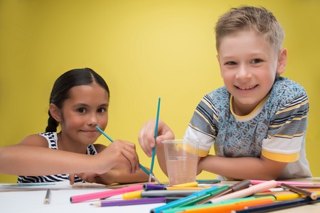 Half-length portrait of little girl wearing nice striped dress sitting at the table with many watercolors and felt-tips on it painting and laughing with little fair-haired boy together. の写真素材