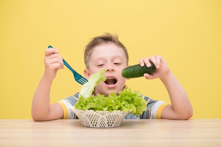 Half-length portrait of lovely little fair-haired funny boy wearing T-shirt sitting at the table wanted to bite the cucumber dawdling in the plateful of lettuceの写真素材