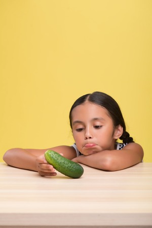 Half-length portrait of pretty dark-haired sad little girl wearing nice striped dress sitting at the table and dolefully looking at the cucumber. Isolated on yellow background の写真素材
