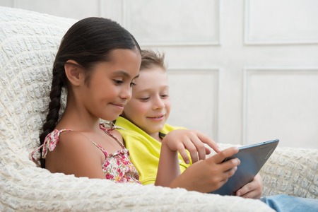 Portrait of two lovely kids sitting aside in the great cozy armchair looking at something on the tablet spend good time togetherの写真素材