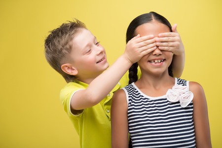 Half-length portrait of lovely little smiling girl wearing nice striped dress and fair-haired smiling boy wearing yellow shirt closing eyes of girl with his hands. Isolated on yellow background の写真素材