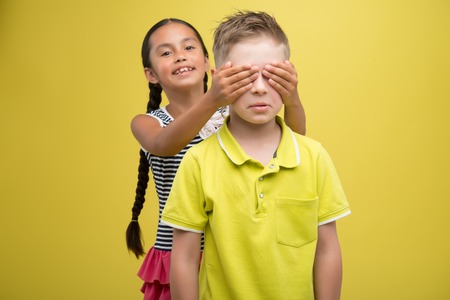 Half-length portrait of lovely little smiling girl wearing nice striped dress closing eyes with his hands of fair-haired sad boy wearing yellow shirt. Isolated on yellow background の写真素材