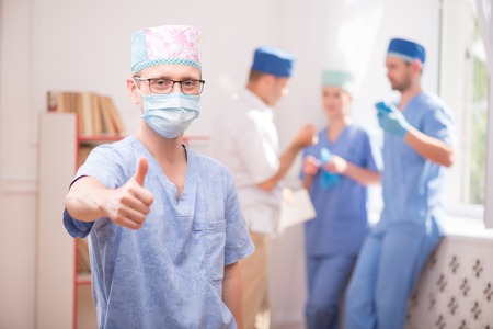 Selective focus on the surgeon wearing blue medical dress showing that everything is OK. His colleagues standing near the window on backgroundの写真素材