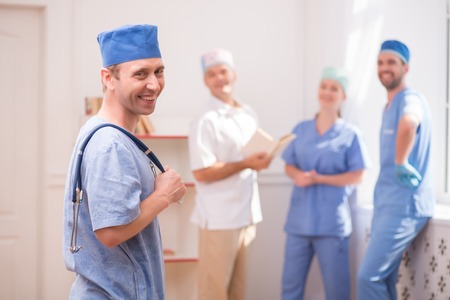 Selective focus on smiling doctor wearing blue medical dress standing aside keeping a stethoscope and looking at us. His smiling colleagues standing near the window on backgroundの写真素材