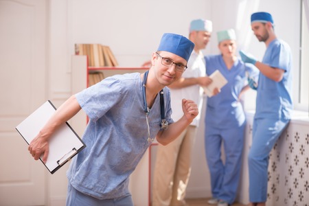 Selective focus on smiling doctor wearing eyeglasses and blue medical dress keeping a stethoscope on his shoulders and holding a folder satisfied with his job. His colleagues discussing some problem on backgroundの写真素材