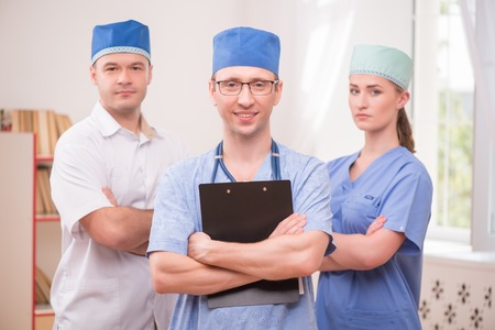 Selective focus on the smiling doctor wearing eyeglasses and blue medical dress keeping a stethoscope on his shoulders and holding a folder. Two his colleagues on backgroundの写真素材