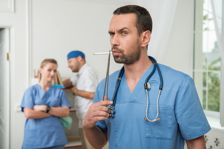 Selective focus on the dark-haired serious surgeon wearing blue medical uniform keeping a stethoscope on his shoulders trying to smoke with the help of tweezers. His colleagues standing on background の写真素材