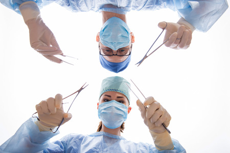 Half-length portrait of two doctors man and woman wearing blue medical masks and uniform holding scissors and tweezers in both hands operating the patient looking at us. Low angle の写真素材