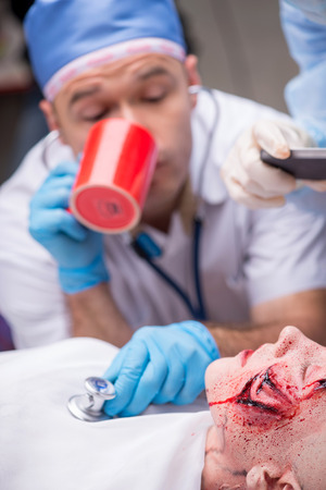 Selective focus on the woman lying on the operation table with the big green wound on her face. Doctor drinking coffee and sounding heart of his patient の写真素材