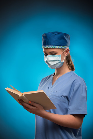Half-length portrait of the fair-haired nurse wearing blue medical uniform and masks standing aside and reading a book. Isolated on blue の写真素材