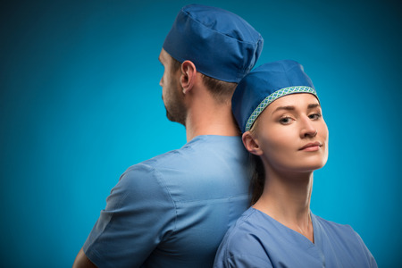 Half-length portrait of two young doctors wearing blue medical uniform standing back to each other...