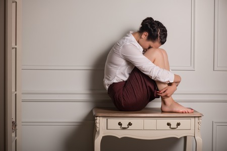 Full-length portrait of beautiful dark-haired thoughtful young woman wearing white blouse and vinous skirt sitting on the little table leaning to the wallの写真素材