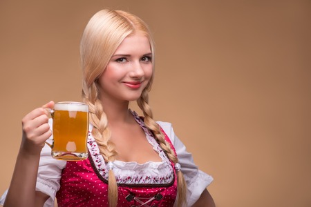 Young sexy smiling blonde wearing pink dirndl with white blouse holding beer mug showing us how she likes it. Isolated on dark backgroundの写真素材