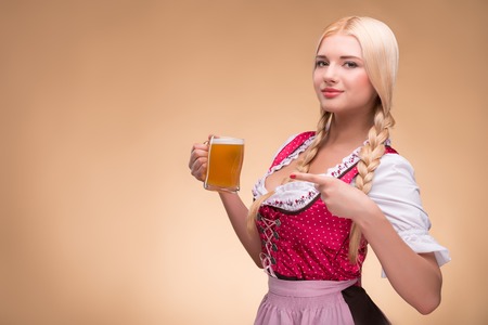 Half-length portrait of young sexy smiling blonde wearing pink dirndl with white blouse looking at us standing aside pointing at the beer mug. Isolated on dark backgroundの写真素材