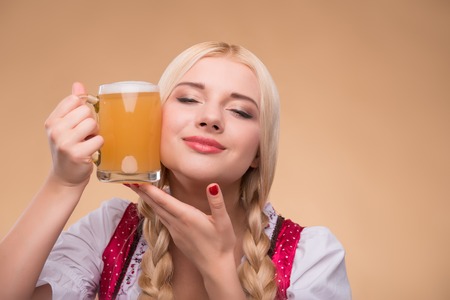 Half-length portrait of young sexy blonde standing with closed eyes wearing pink dirndl with white blouse leaning to the mug of beer. Isolated on dark backgroundの写真素材