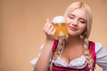 Half-length portrait of young sexy blonde standing with closed eyes wearing pink dirndl with white blouse leaning to the mug of beer. Isolated on dark backgroundの写真素材