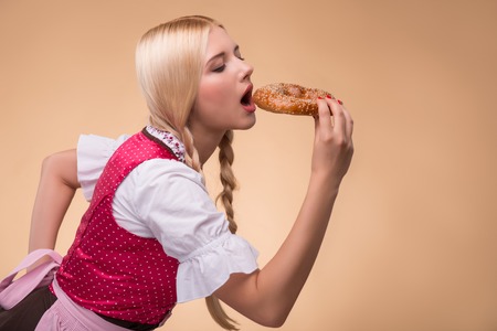 Half-length portrait of young sexy blonde standing aside with closed eyes wearing pink dirndl and white blouse tasting very delicious bagel. Isolated on dark backgroundの写真素材