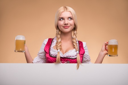 Half-length portrait of young sexy smiling blonde wearing pink dirndl and white blouse holding two beer mugs looking out of the big white poster for copy place. Isolated on dark backgroundの写真素材