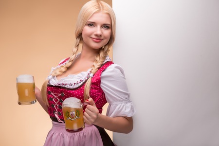 Half-length portrait of young sexy smiling blonde wearing pink dirndl and white blouse holding two beer mugs leaning on the big white poster for copy place. Isolated on dark backgroundの写真素材