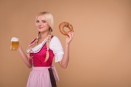 Half-length portrait of young sexy blonde wearing pink dirndl and white blouse standing aside holding a bagel in one hand and beer mug in another. Isolated on dark backgroundの写真素材