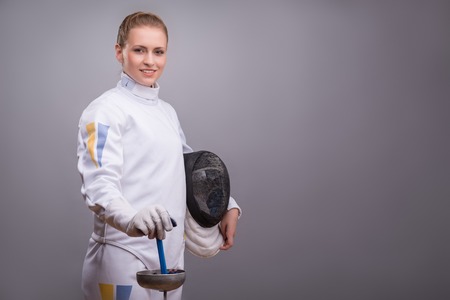 Half-length portrait of the fair-haired smiling girl wearing white fencing costume standing aside holding the fencing mask in one hand and a rapier in another. の写真素材