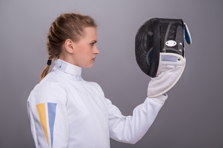 Half-length portrait of pretty serious girl wearing fencing costume standing aside looking at her fencing mask. Isolated on dark backgroundの写真素材