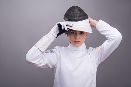Half-length portrait of the fair-haired serious girl wearing white fencing costume putting on her fencing mask. Isolated on grey backgroundの写真素材