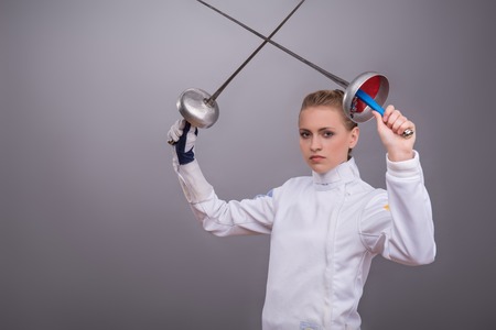 Half-length portrait of the fair-haired pretty serious girl wearing white fencing costume standing crossing two swords and looking at us. Isolated on grey backgroundの写真素材