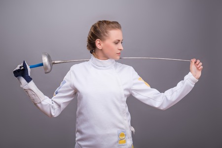 Half-length portrait of pretty smiling girl wearing fencing costume holding her sword on the shoulders thinking about future competitions. Isolated on dark backgroundの写真素材