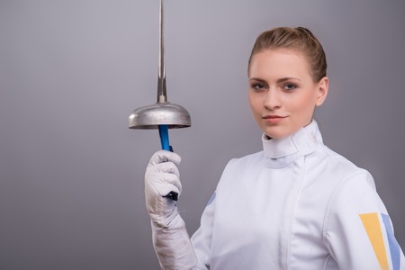 Half-length portrait of the fair-haired pretty girl wearing white fencing costume standing aside presenting us her sward. Isolated on grey backgroundの写真素材