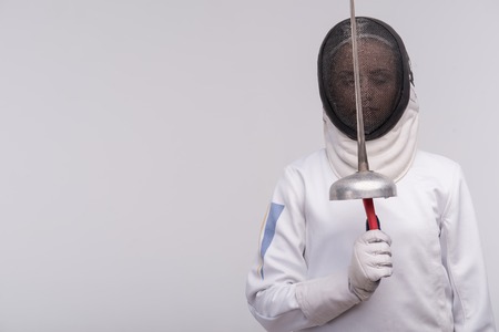 Half-length portrait of woman wearing white fencing costume and black fencing mask holding her sword behind her. Isolated on white backgroundの写真素材