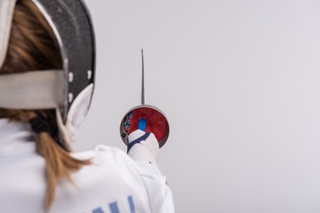 Selective focus on the sword in the hands of woman wearing white fencing costume and black fencing...