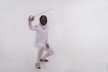Full-length portrait of woman wearing white fencing costume and black fencing mask sitting...