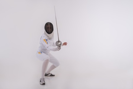 Full-length portrait of woman wearing white fencing costume and black fencing mask sitting practicing with the sword and looking at us. Isolated on white backgroundの写真素材