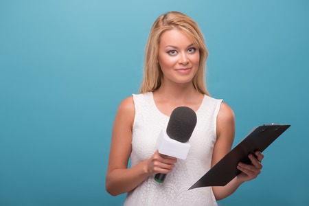 Half-length portrait of lovely fair-haired TV presenter wearing pretty white dress standing to us holding a microphone holding a folder. Isolated on blue backgroundの写真素材