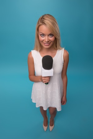 Half-length portrait of lovely smiling fair-haired TV presenter wearing pretty white dress holding a microphone. Top view. Isolated on blue backgroundの写真素材