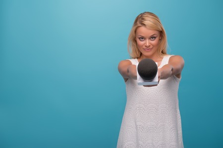 Half-length portrait of lovely smiling fair-haired TV presenter wearing pretty white dress holding a microphone wanted to interview everybody. Isolated on blue backgroundの写真素材