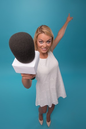 Half-length portrait of lovely smiling fair-haired TV presenter wearing pretty white dress holding a microphone wanted to interview us. Top view. Isolated on blue backgroundの写真素材