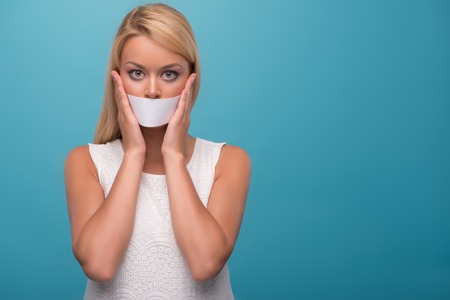 Half-length portrait of lovely fair-haired TV presenter wearing pretty white dress closing her mouth with Scotch tape. Isolated on blue backgroundの写真素材
