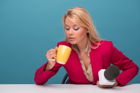 Half-length portrait of lovely fair-haired TV presenter wearing great red jacket and cream-colored shirt sitting at the table looking at cup of coffee. Isolated on blue backgroundの写真素材