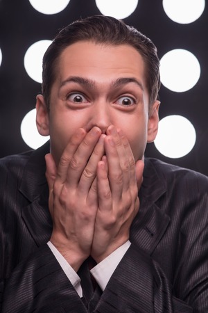 Half- length portrait of handsome astonished man wearing great black suit and vinous tie standing behind the rostrum delighted with the news he has just heard closing his mouth not to screamの写真素材
