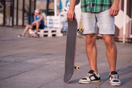 Selective focus on the young man wearing shorts checked T-shirt and trainers standing with his skateboard on the streetの写真素材