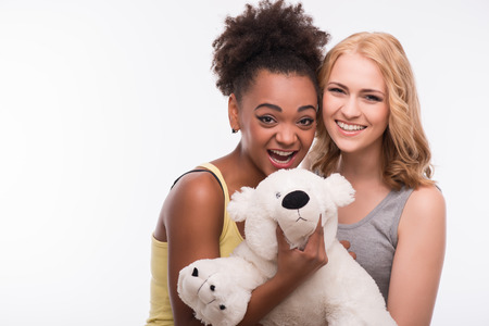 Half-length portrait of two young pretty smiling women wearing colorful T-shirt standing with a white teddy-bear showing that everything is great. Isolated on white backgroundの写真素材