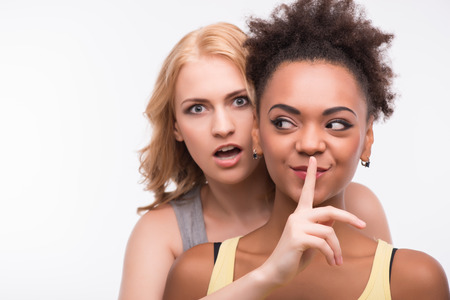 Half-length portrait of young blonde wearing colorful T-shirt hiding behind her smiling friend asking her to be silent looking at us with surprise. Isolated on white backgroundの写真素材
