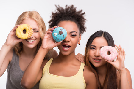 Half-length portrait of three beautiful tempting girls wearing colorful T-shirts having fun looking through the bright delicious doughnuts. Isolated on white backgroundの写真素材